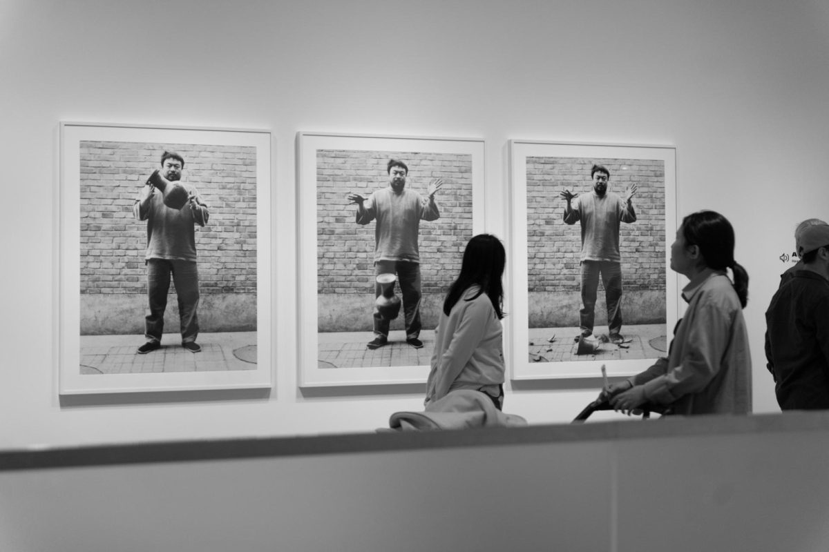 Three people in a gallery observe three large, black-and-white portraits by Ai Weiwei, each capturing a moment of dropping a ceramic object—a striking example of contemporary Chinese art. The gallery wall and viewers are shown in grayscale at SAM.
