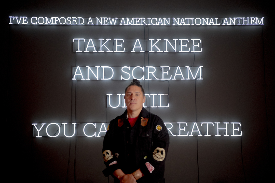 A person stands before a neon art installation by Nicholas Galanin at the Seattle Art Museum. It reads: IVE COMPOSED A NEW AMERICAN NATIONAL ANTHEM: TAKE A KNEE AND SCREAM UNTIL YOU CAN BREATHE. They wear a jacket adorned with patches, embodying unconventional patriotism.