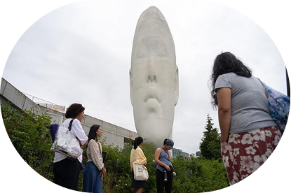 People standing and observing a large, white, sculpture of an elongated head set against an overcast sky, surrounded by greenery. 
