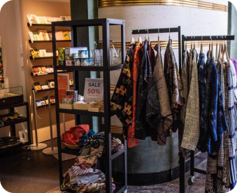 A boutique shop featuring a clothing rack with colorful garments, a shelf displaying a Sale 50% sign, and various accessories. Display shelves with books and cards are in the background.