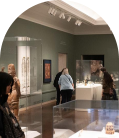 A museum gallery with visitors observing artifacts displayed in glass cases. The room features green walls and various sculptures and art pieces. 