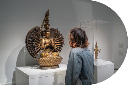 A person in a denim jacket observes a multi-armed statue of a seated deity displayed on a pedestal in a museum. 