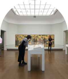 A person closely examines a display case in an art gallery. Behind them, another person observes a large artwork depicting an outdoor scene. The room features modern lighting and a polished wooden floor.