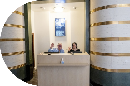 Two Seattle Art Museum staff waving at the camera sitting behind a desk situated between two ornate columns..
