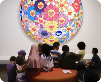a group of children sit in front of a large, circlular artwork covered in colorful flowers.