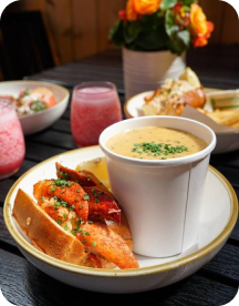 a close up shot of a plate with a lobster roll and clam chowder, in the background a few pinky drinks in short glasses