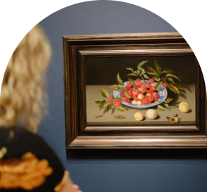 A person observes a framed still-life painting of a blue bowl filled with fruit and adorned with leaves, placed on a wooden table.