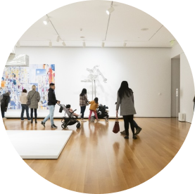 A group of people, including some with strollers, walk through a modern art gallery with wooden floors. Colorful abstract paintings and sculptures are displayed.