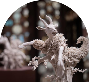 A sculpture of a goat with an ornate, textured body resembling coral. The goat is poised with dynamic movement on a pedestal, captured in soft lighting.