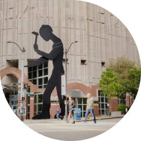 A large silhouette sculpture of a person hammering in front of the Seattle Art Museum building. People walk past on the sidewalk, and trees are visible nearby.