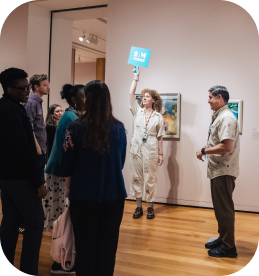A museum guide holds up a blue sign, leading a group through the Seattle Art Museum’s exhibitions, with framed paintings on the walls and wooden floors.