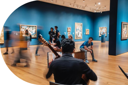 A person plays guitar in the foreground of an art gallery with blue walls, while people walk around and observe paintings in the background. Some visitors are seated on benches.