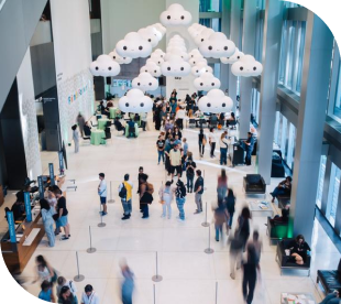 A busy, modern indoor lobby with people walking and gathering—much like the vibrant atmosphere near Pike Place Market. Large, cloud-shaped white light fixtures hang from the high ceiling above the crowd. Tall windows line one side, letting in natural light.